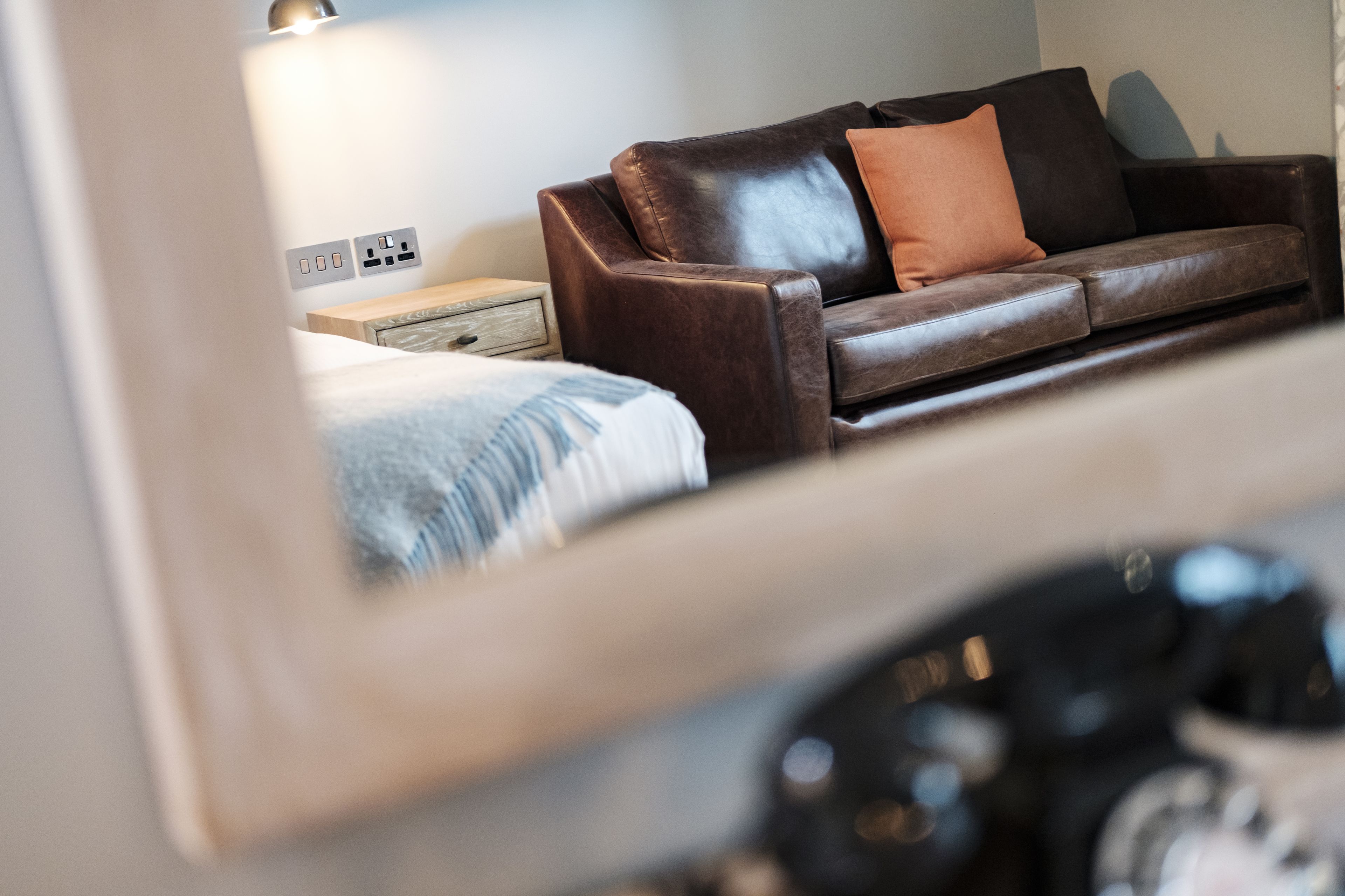A room with a brown leather sofa, a wooden nightstand, and a bed with a blue blanket.