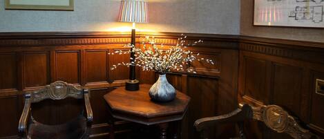 A corner room with a wooden paneled wall, a table with a vase of flowers, two chairs, a lamp, and a framed architectural drawing.