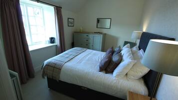 A bedroom with a bed, a bedside table with a lamp, a dresser, a window with curtains, and a wall-mounted mirror.