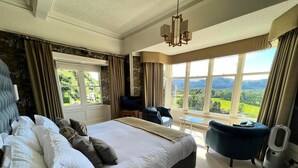 A spacious bedroom with a large bed, a sitting area with a chair, a TV, and a view of the outdoors. - Ravenstone Manor (Keswick)