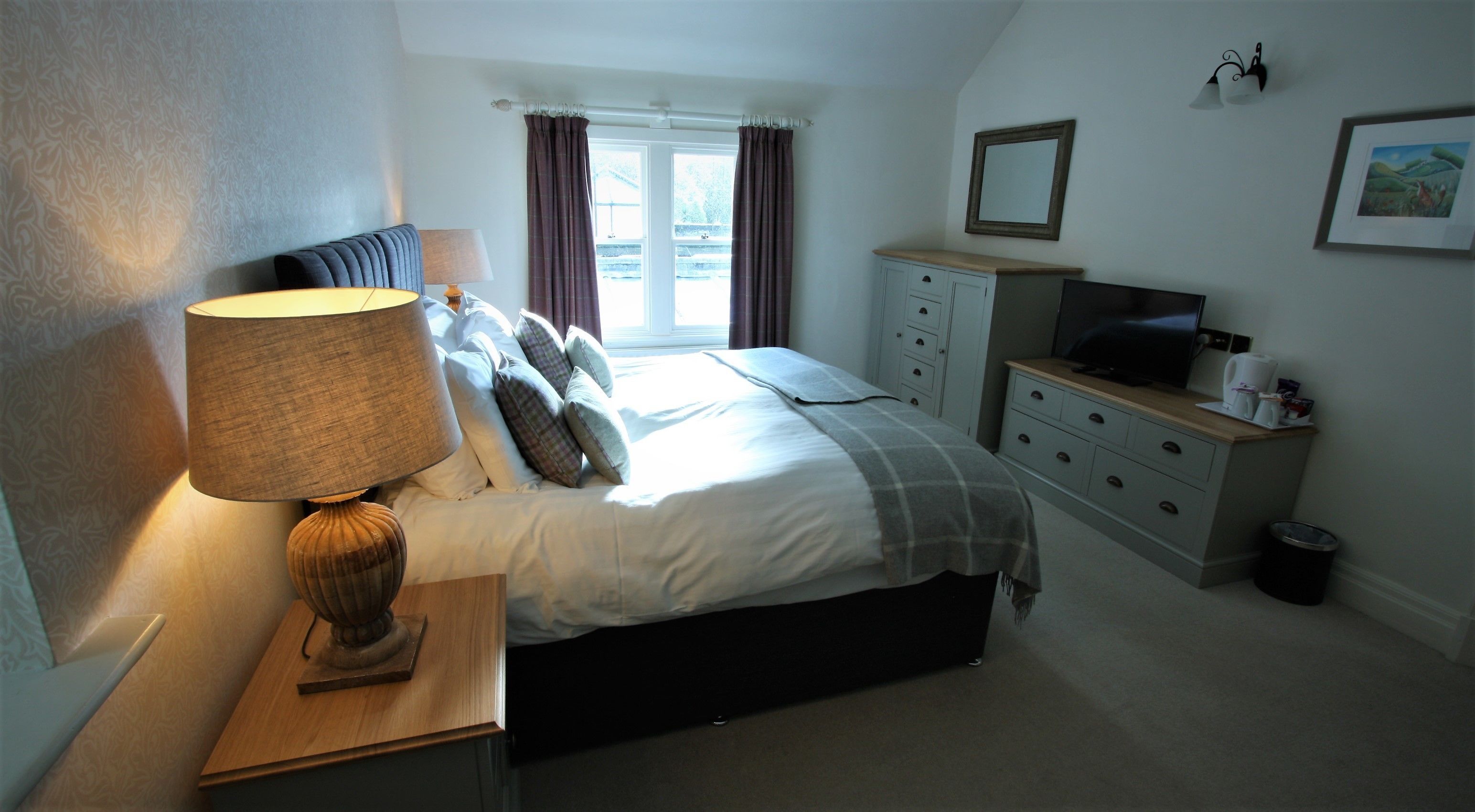 A bedroom with a bed, bedside lamp, dresser, television, and a window with curtains.