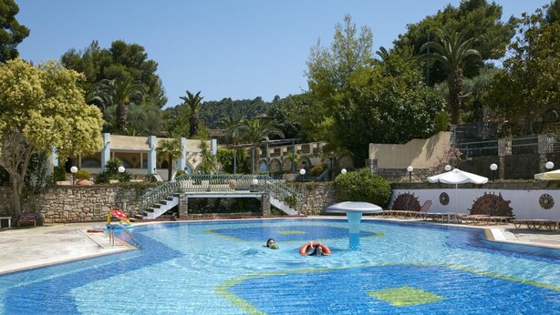 A swimming pool with a tiled design, surrounded by lounge chairs and umbrellas, with a stone wall and trees in the background. - GHotels Macedonian Sun (Kassandra)