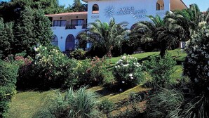 A two-story building with a white facade and red-tiled roof, surrounded by lush greenery and palm trees. - GHotels Macedonian Sun (Kassandra)