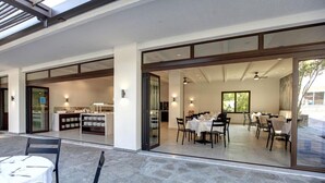 An outdoor dining area with tables and chairs, a kitchen visible through glass doors, and a large window providing a view of the outdoors. - GHotels Macedonian Sun (Kassandra)