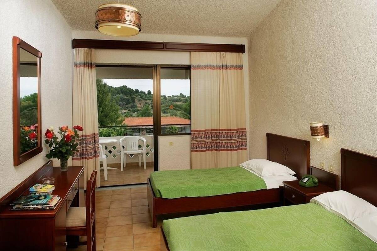 A hotel room with two beds, a desk with a chair, a small table with a chair, a balcony with a view of greenery and buildings, and a telephone on the bedside table.