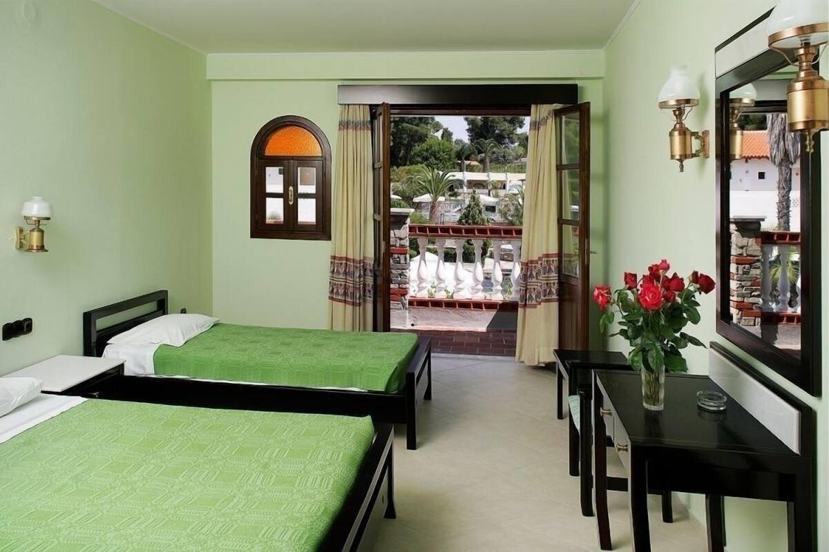 A hotel room with two beds, a balcony, a dining table, and a vase with flowers.