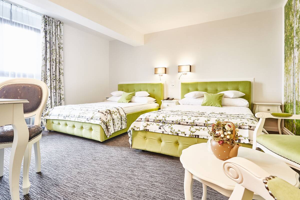 Ein Hotelzimmer mit zwei Betten, einem Schreibtisch, einem Stuhl und Blick auf Bäume durch das Fenster.
