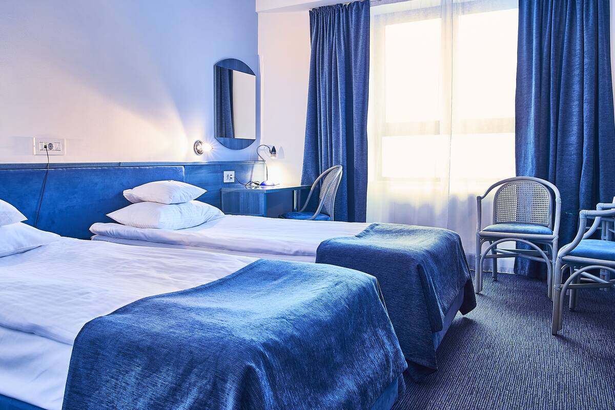 Ein Hotelzimmer mit zwei Betten, einem Schreibtisch, einem Stuhl und einem großen Fenster mit Vorhängen.