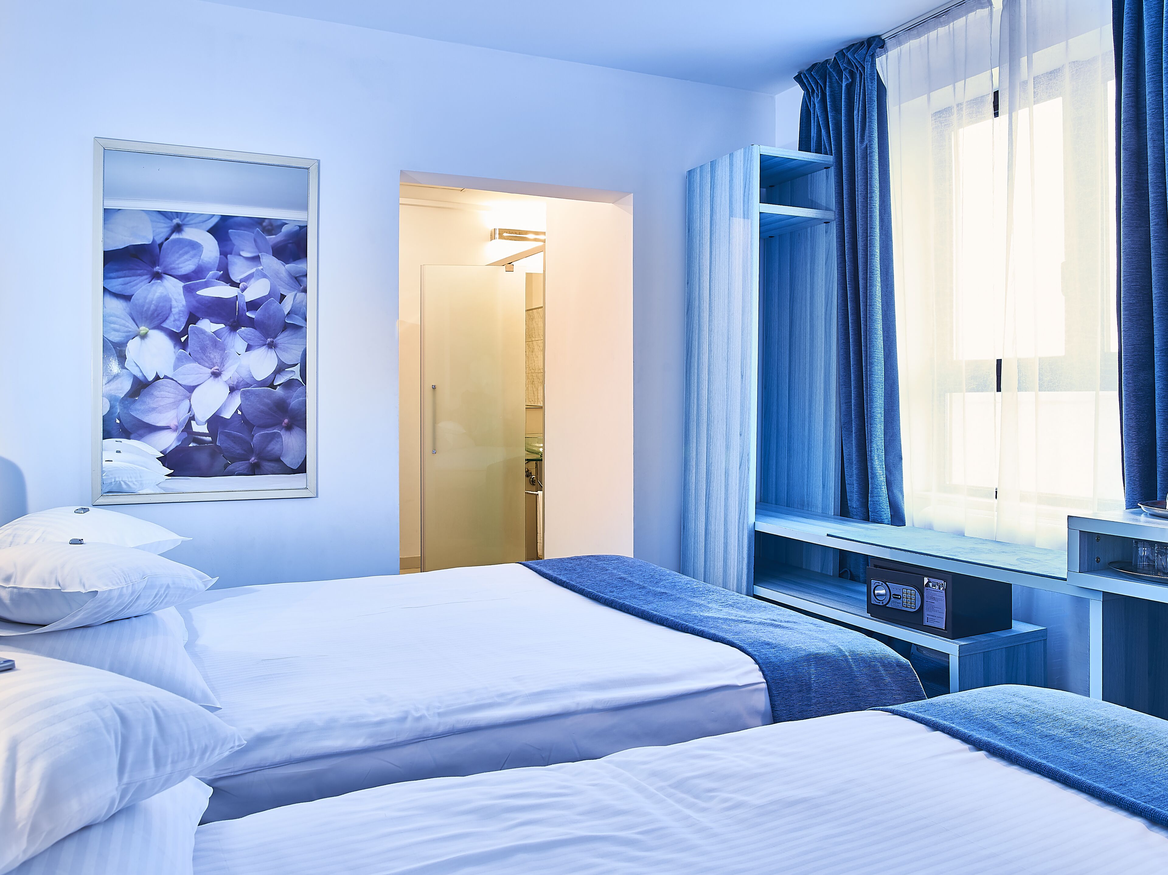 A hotel room with two beds, a large window with curtains, a TV, and a bathroom visible through an open door.