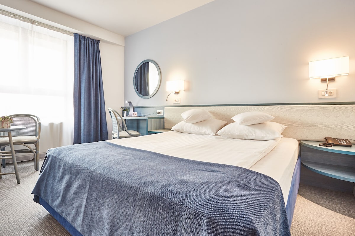 Ein Hotelzimmer mit einem großen Bett, einem Nachttisch mit Lampe, einem Sessel und einem fenster mit Vorhängen.