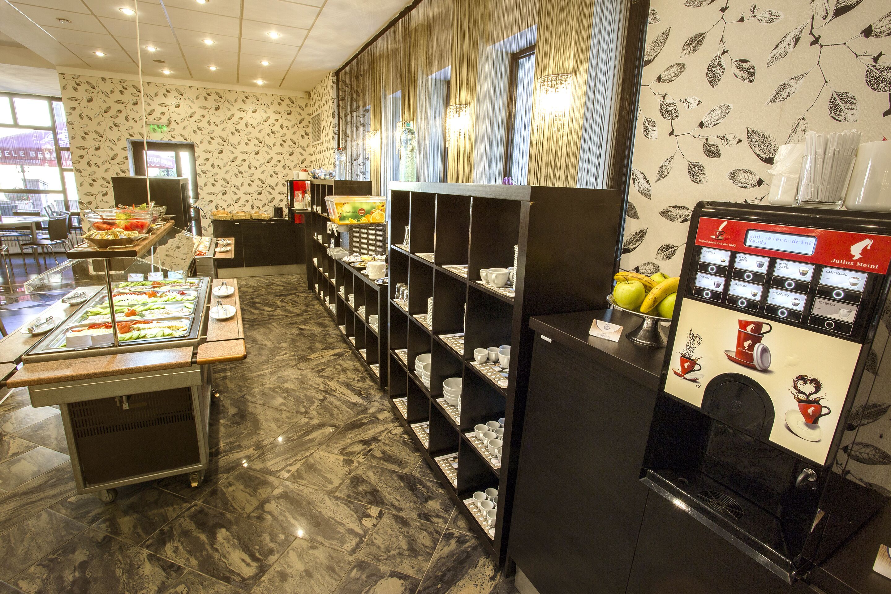 Un espace buffet comprenant une machine à café, un présentoir de fruits et une variété de plats disposés sur des étagères.