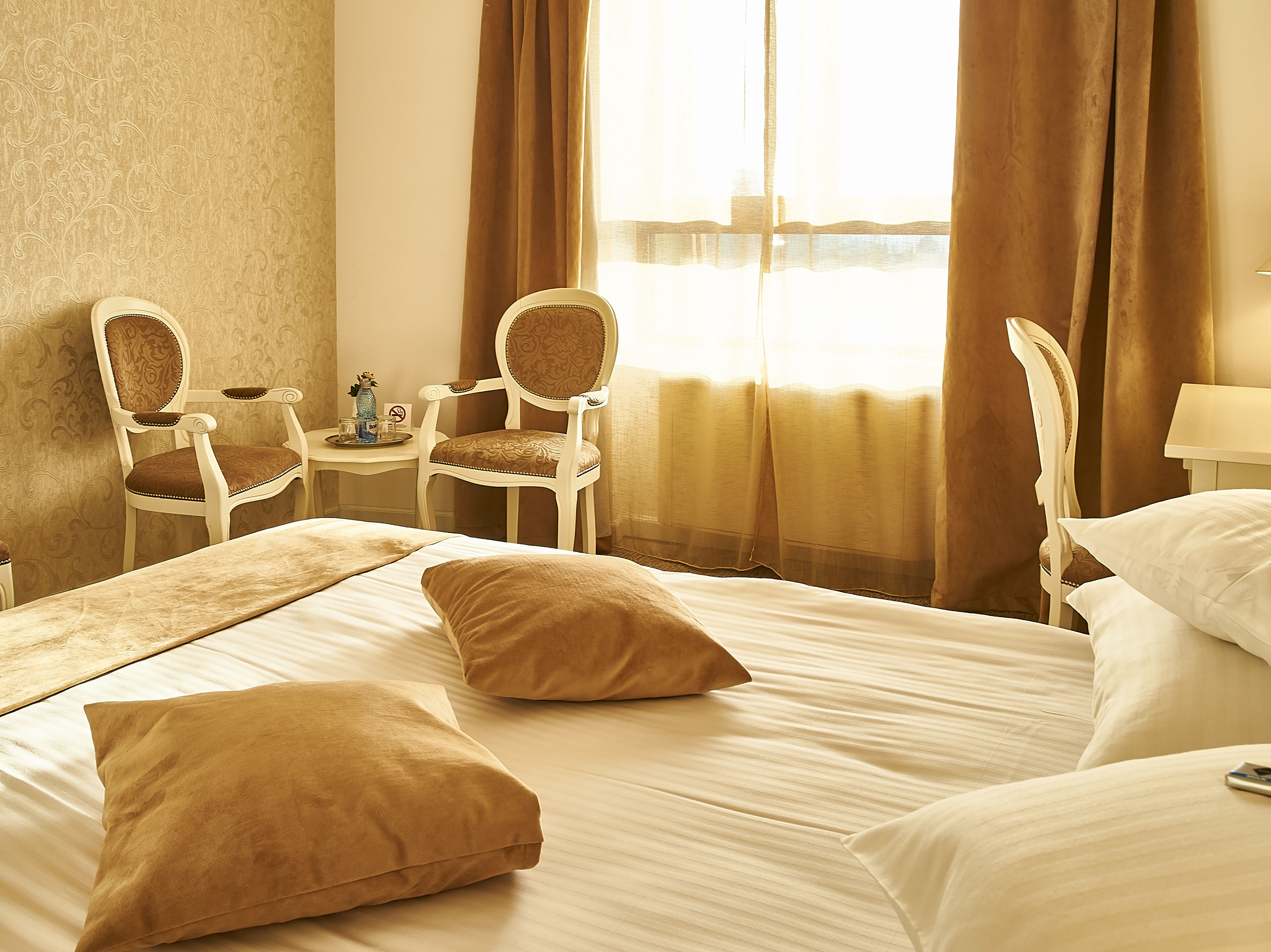Habitación de hotel con una cama grande, dos sillas, una mesita con un florero y vistas al mar a través de cortinas traslúcidas.