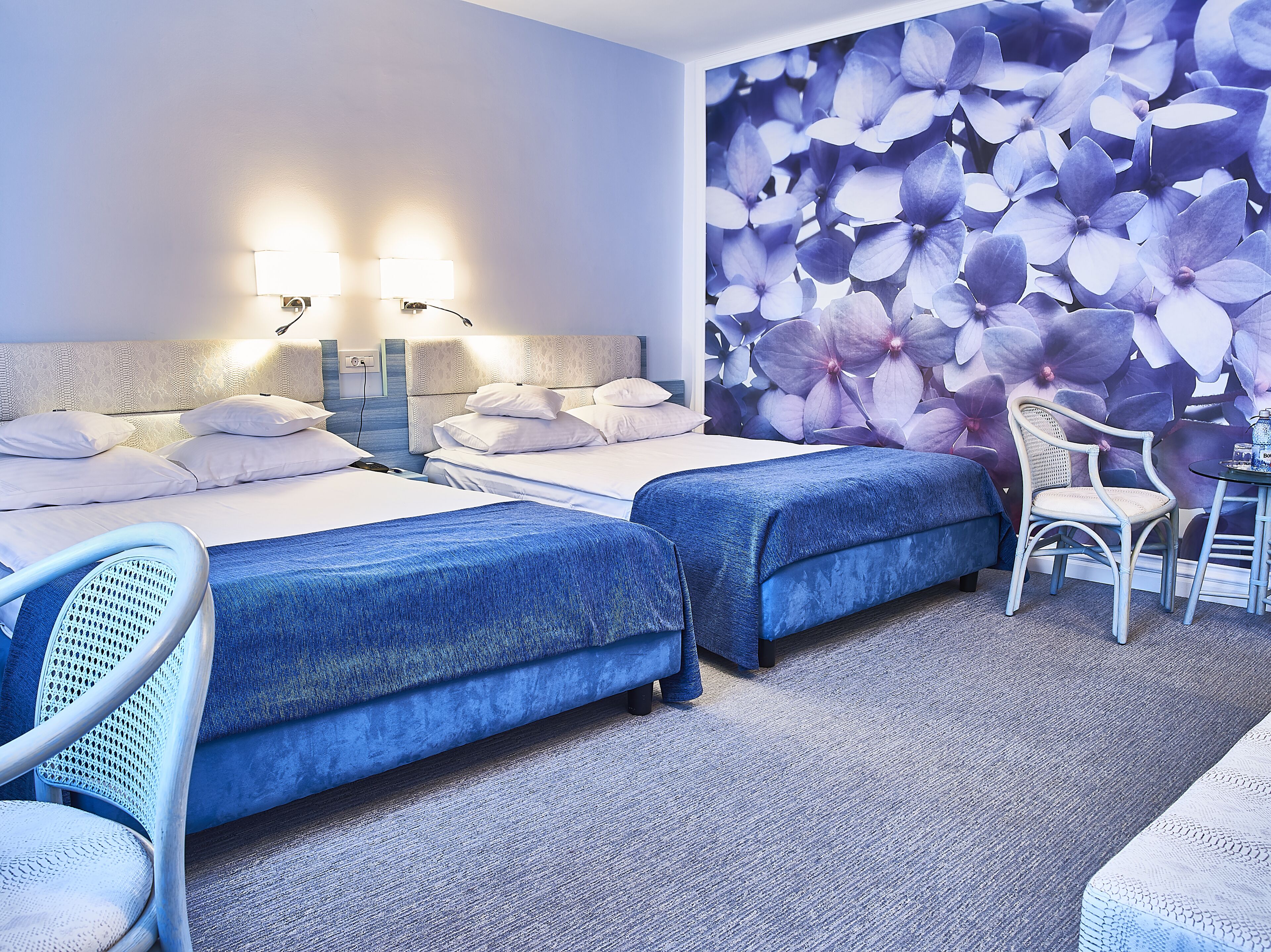 Habitación de hotel con dos camas, una silla, una mesa y una pared con un mural floral.