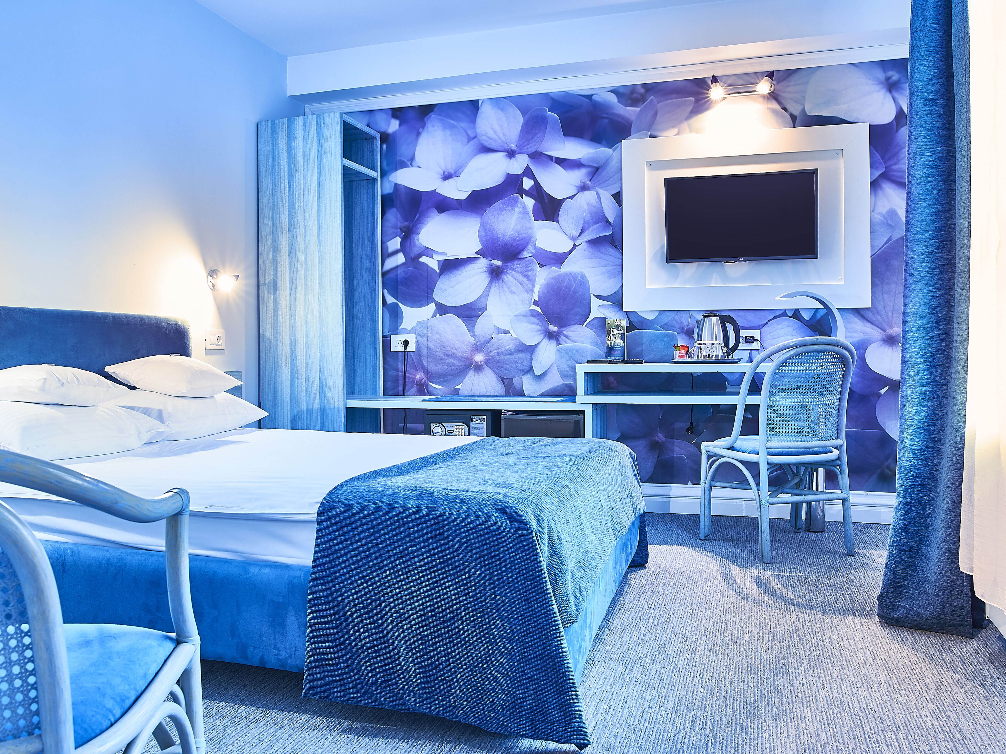 A hotel room with a bed, a desk, a chair, a TV mounted on the wall, and a large floral wallpaper.