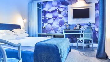 A hotel room with a bed, a desk, a chair, a TV mounted on the wall, and a large floral wallpaper.