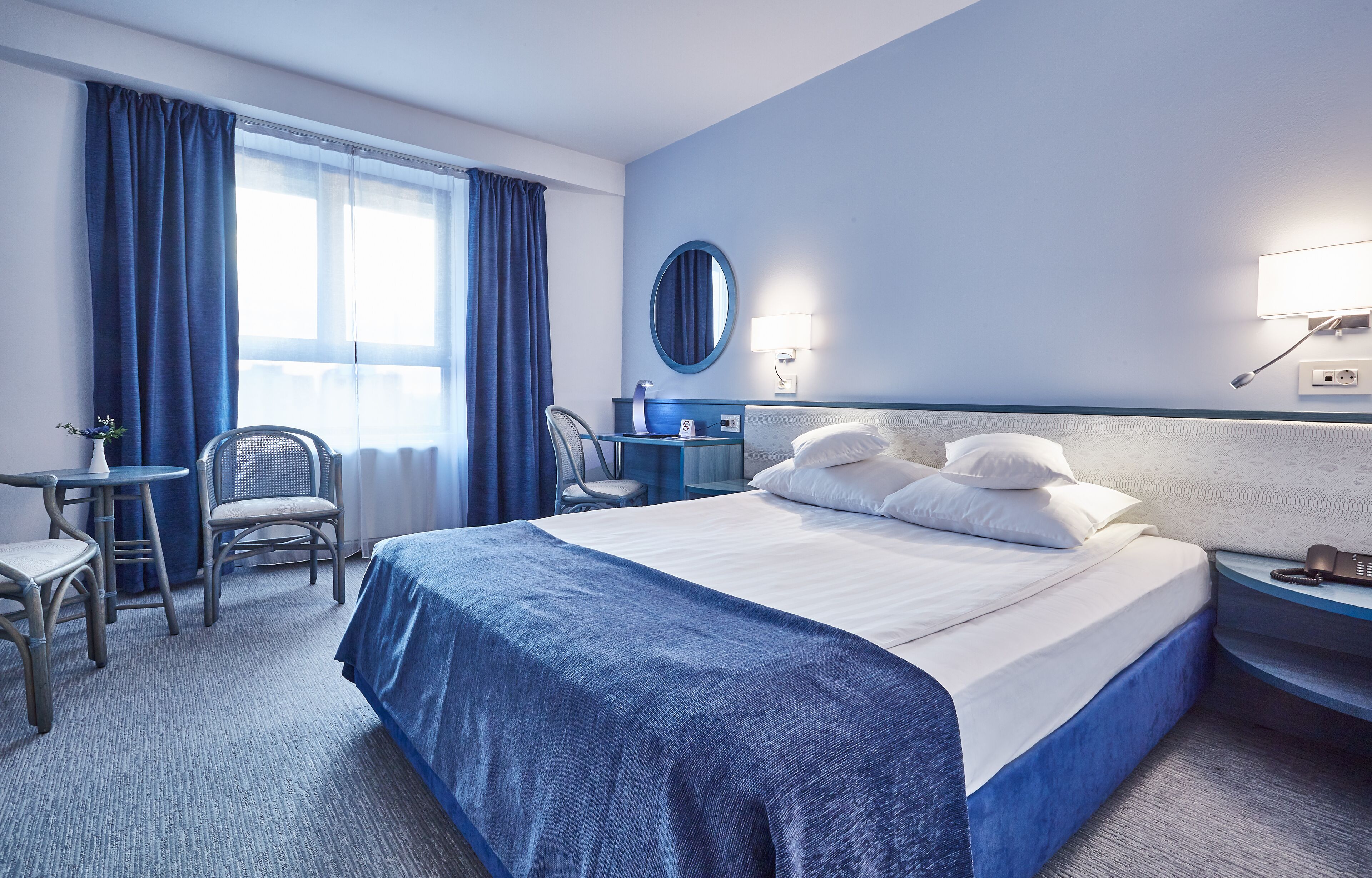 Habitación de hotel con una cama grande, un escritorio con silla, un espejo redondo y una mesita de noche con teléfono.