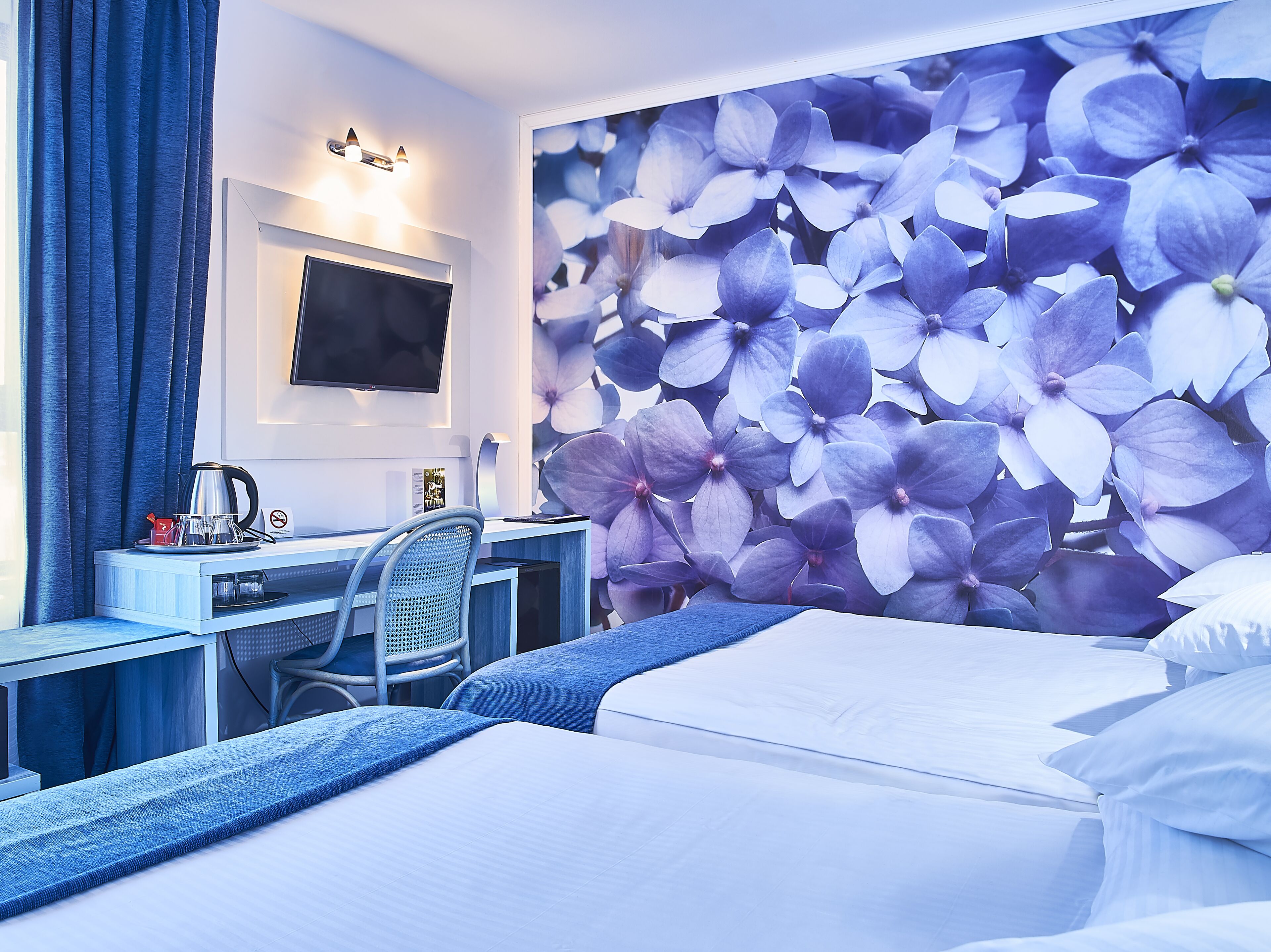 A hotel room with a bed, a desk with a chair, a TV, and a wall with a floral wallpaper.