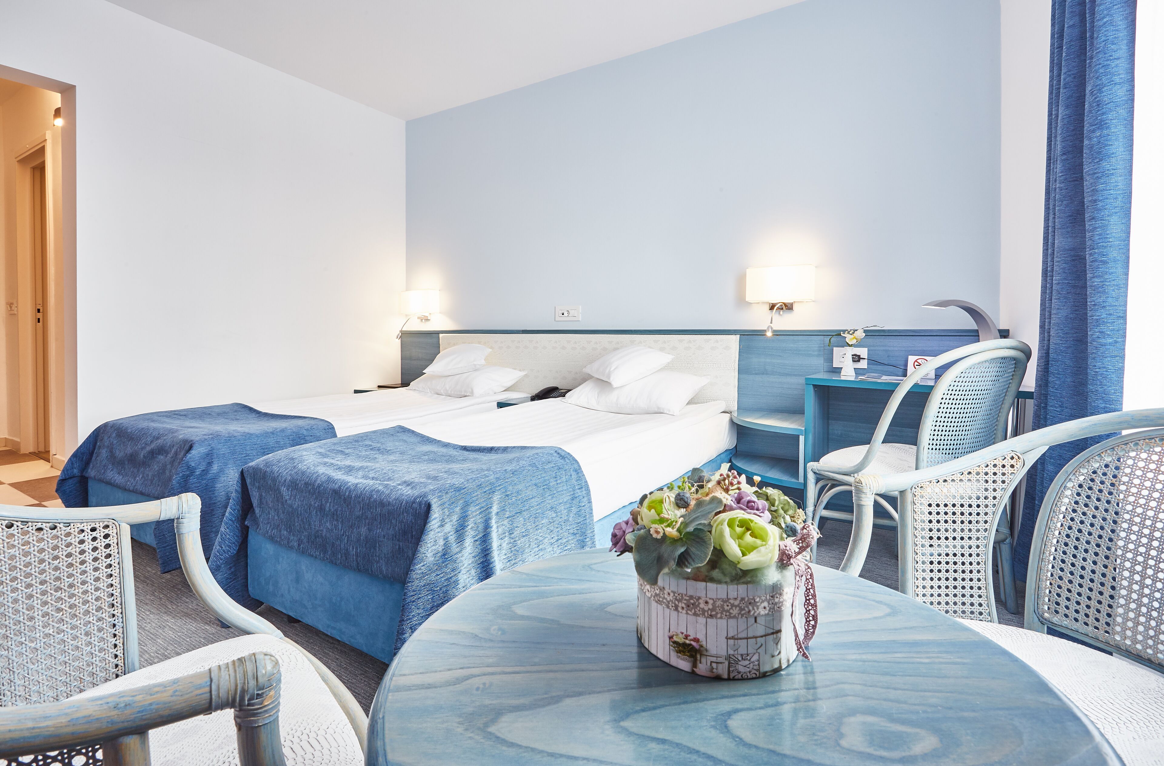 Une chambre d’hôtel avec deux lits, une petite table ornée d’un vase de fleurs et un coin salon avec des fauteuils.