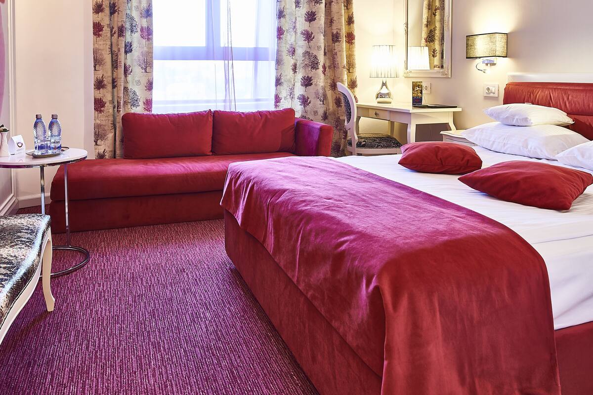 Ein Hotelzimmer mit rotem Sofa, einem Bett mit roter Bettwäsche, einem kleinen Tisch mit Wasserflaschen und einer Lampe.