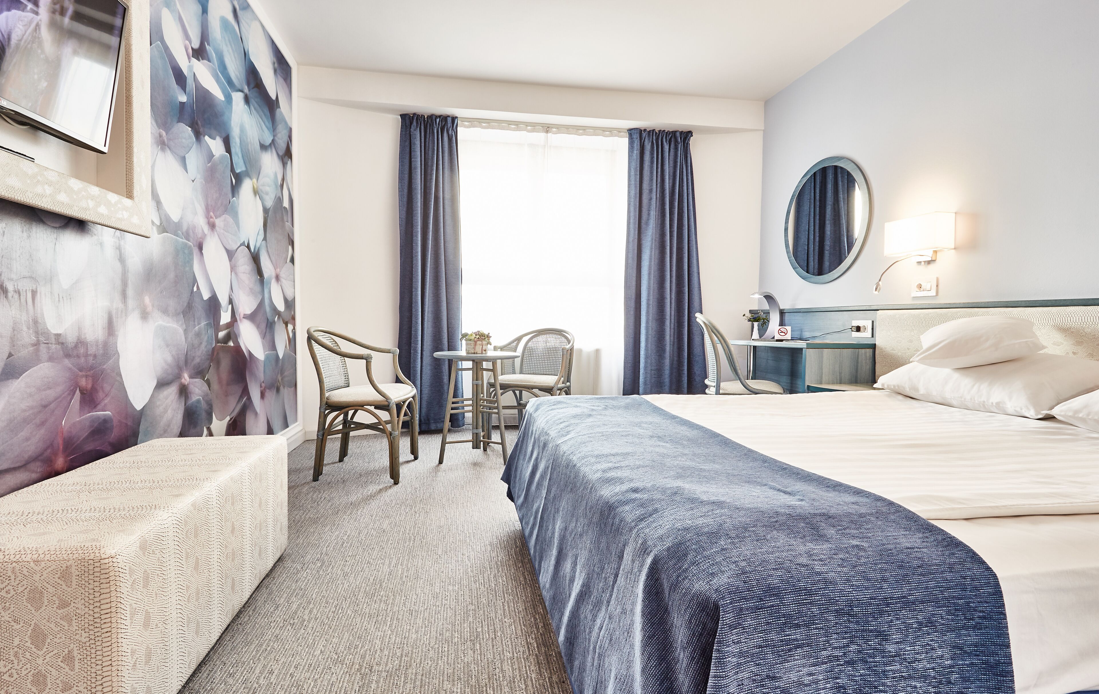 A hotel room with a bed, two chairs, a desk, a mirror, and a window with curtains.