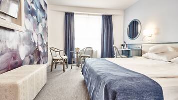 A hotel room with a bed, two chairs, a desk, a mirror, and a window with curtains.