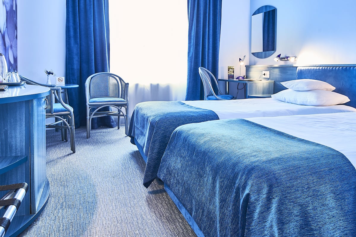 Ein Hotelzimmer mit zwei Betten, einem Schreibtisch, einem Stuhl, einer Kommode und einem großen Fenster mit Vorhängen.