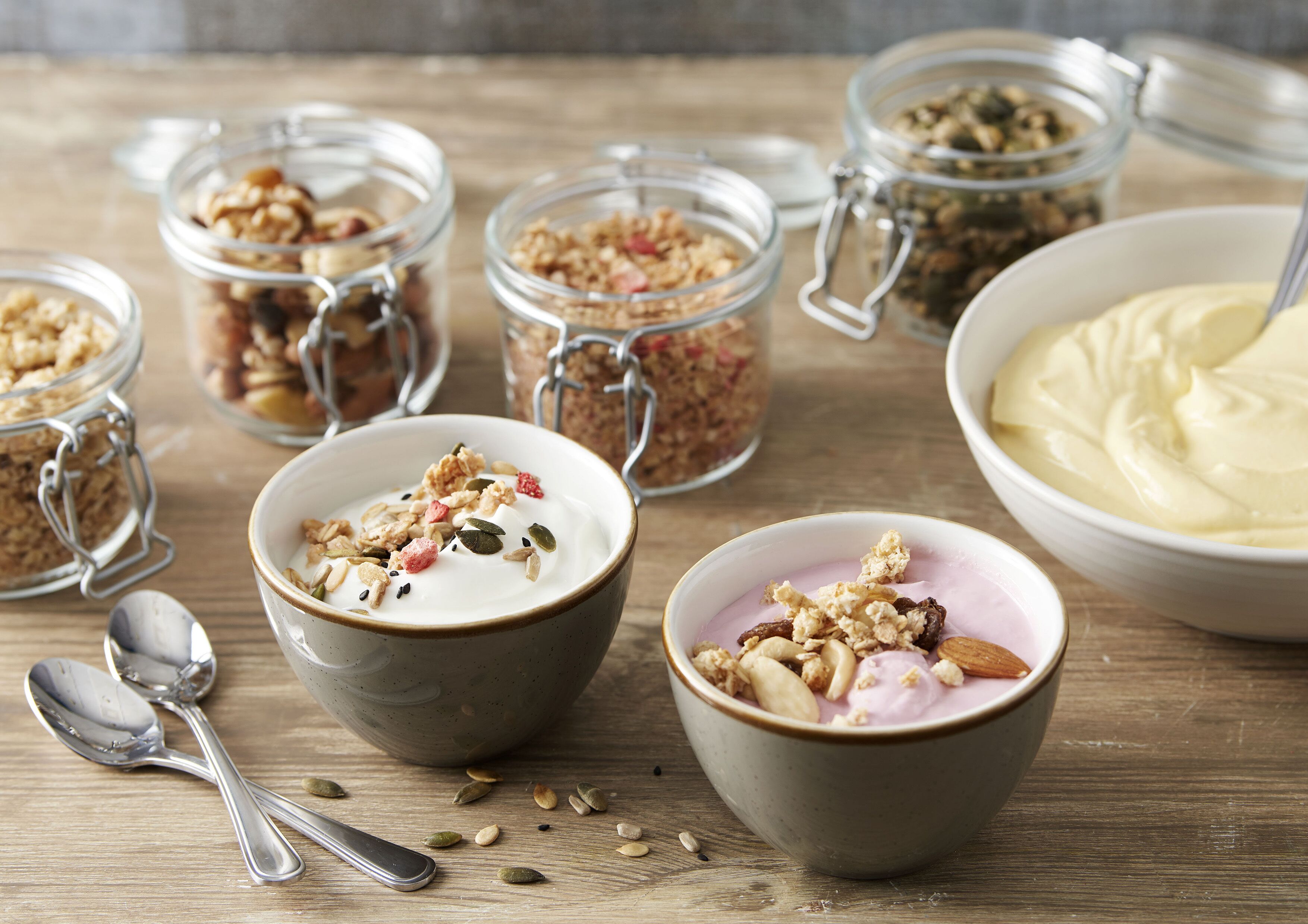 Skåle med yoghurt, granola og frø, samt glas med granola og nødder på et træbord.