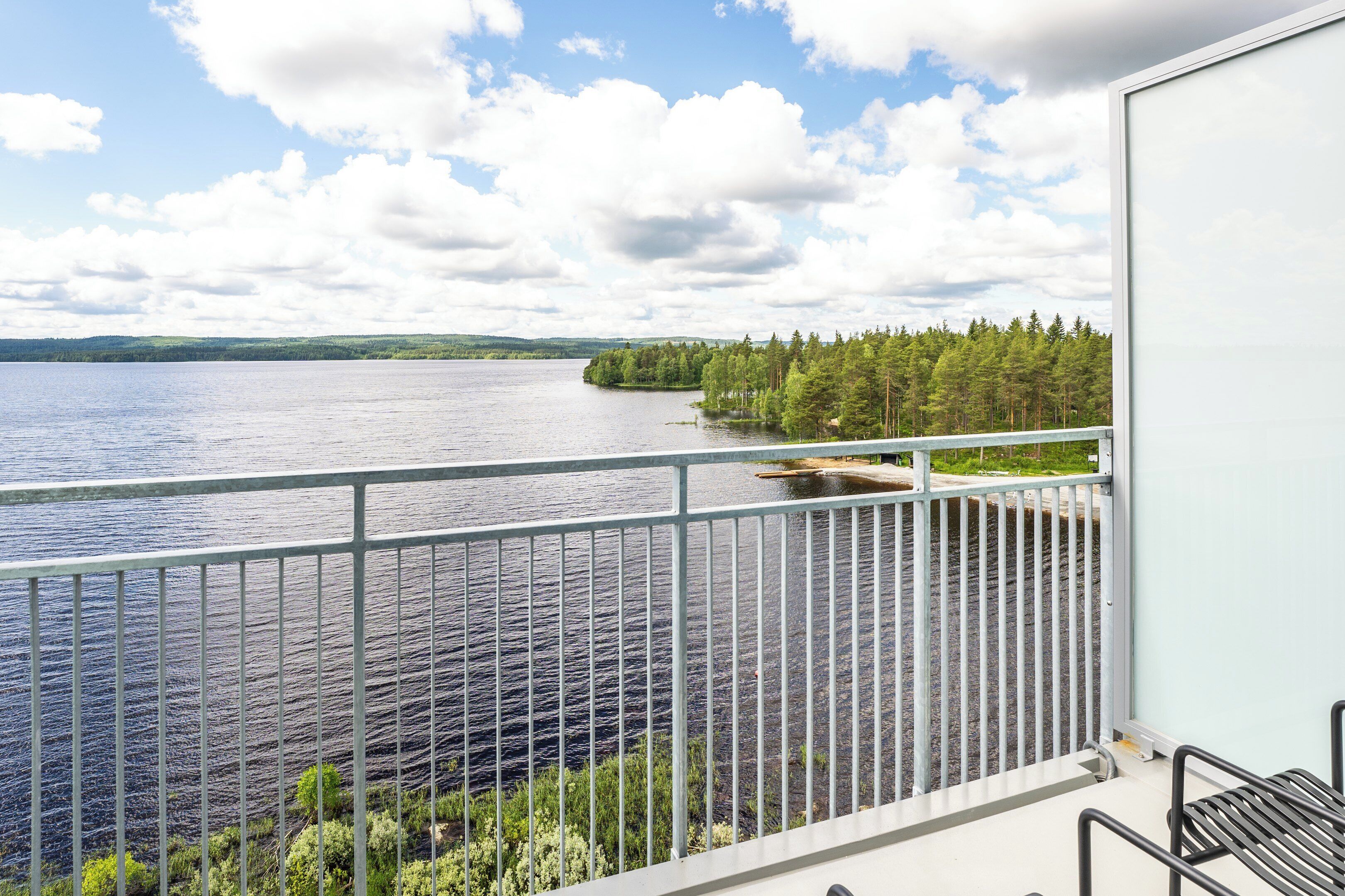 A balcony with a metal railing overlooking a lake, surrounded by trees and a clear sky.