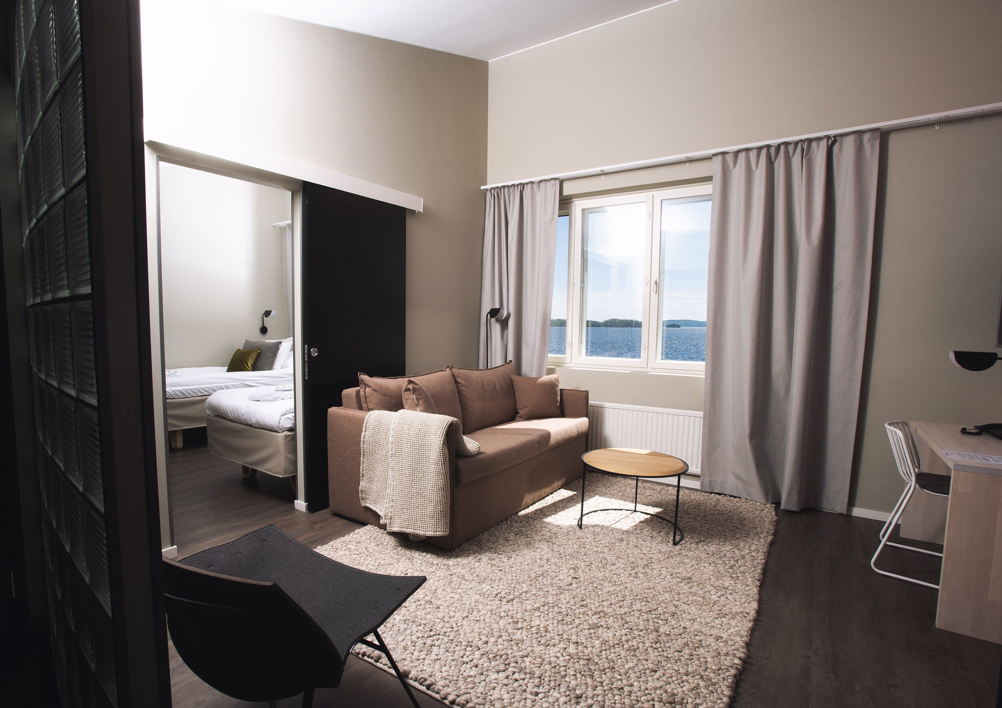 Una habitación moderna y luminosa con un sofá, una mesita y una silla. Se ve una cama al fondo, y una ventana ofrece vistas al exterior.