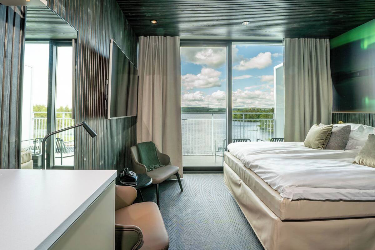 A modern hotel room with a large bed, a desk, a chair, and a balcony with a view of the water.