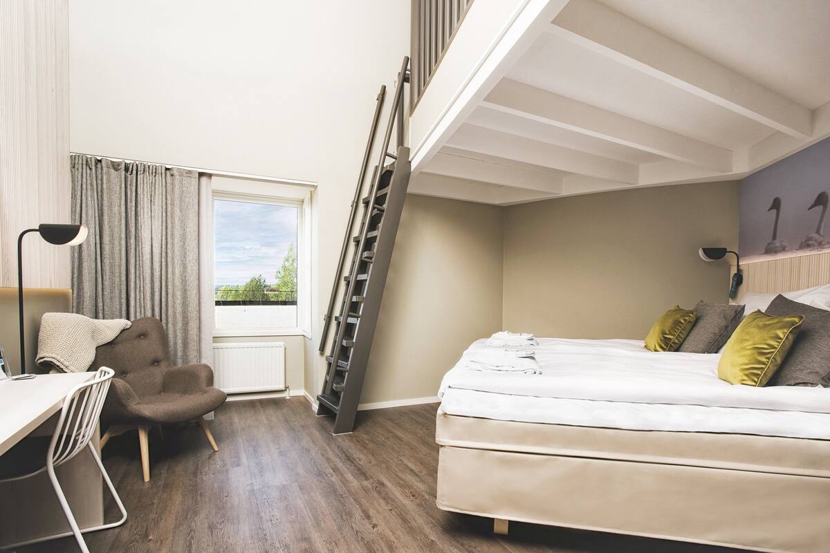 A bedroom with a loft bed, a desk, a chair, and a window with curtains.