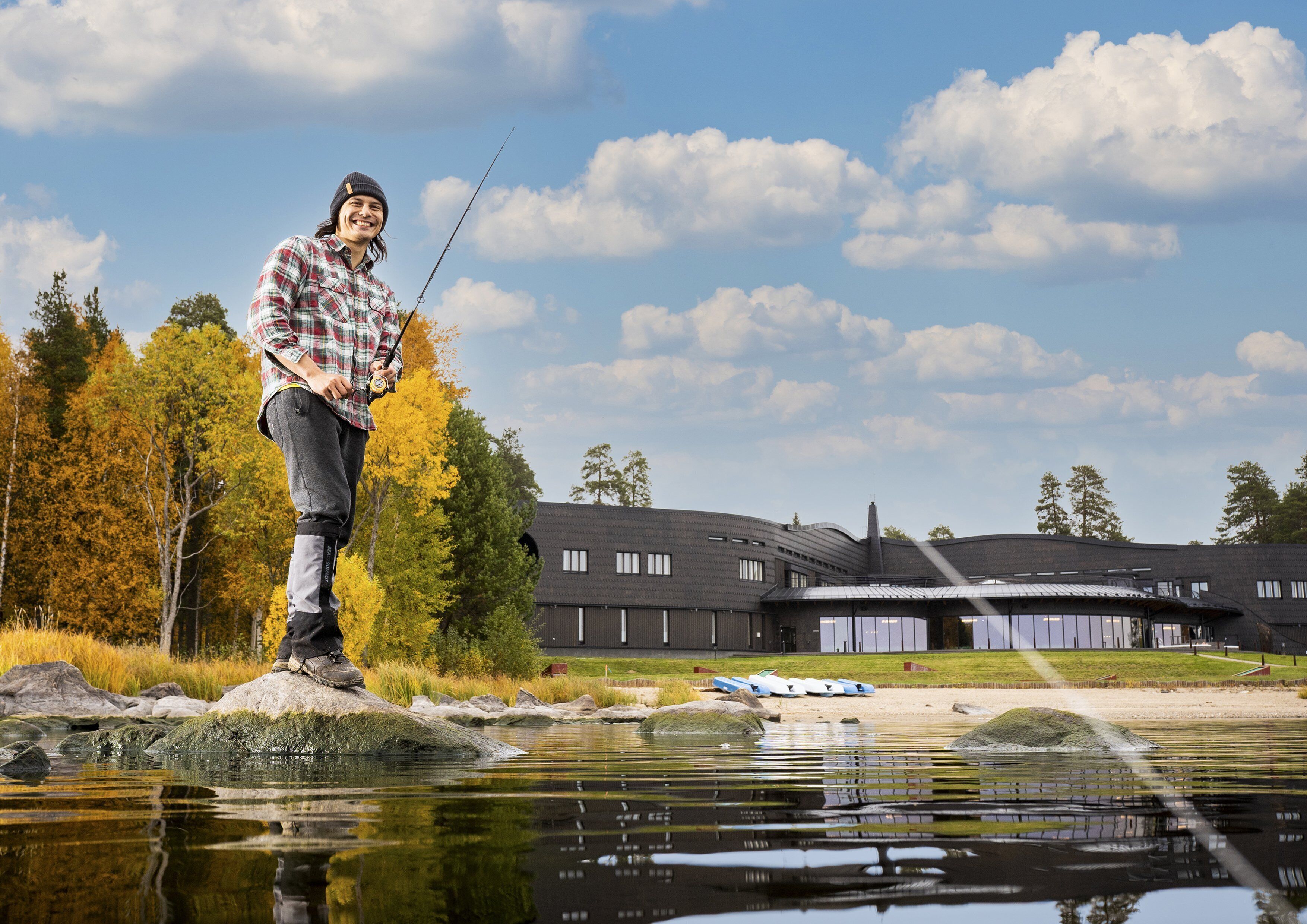 A man fishing by a calm river with a modern building in the background.