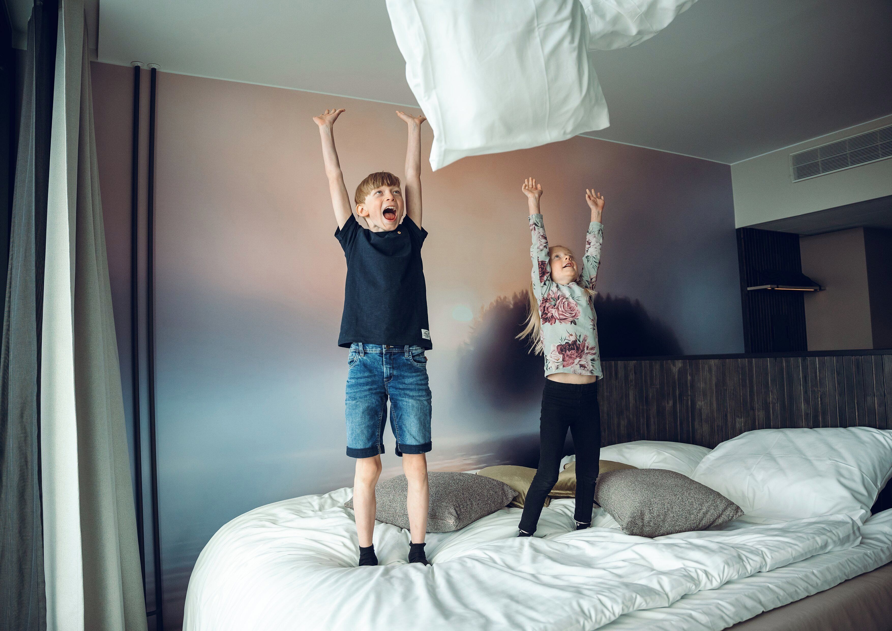 Two children jumping on a bed in a hotel room with a plain wall and a window with curtains.