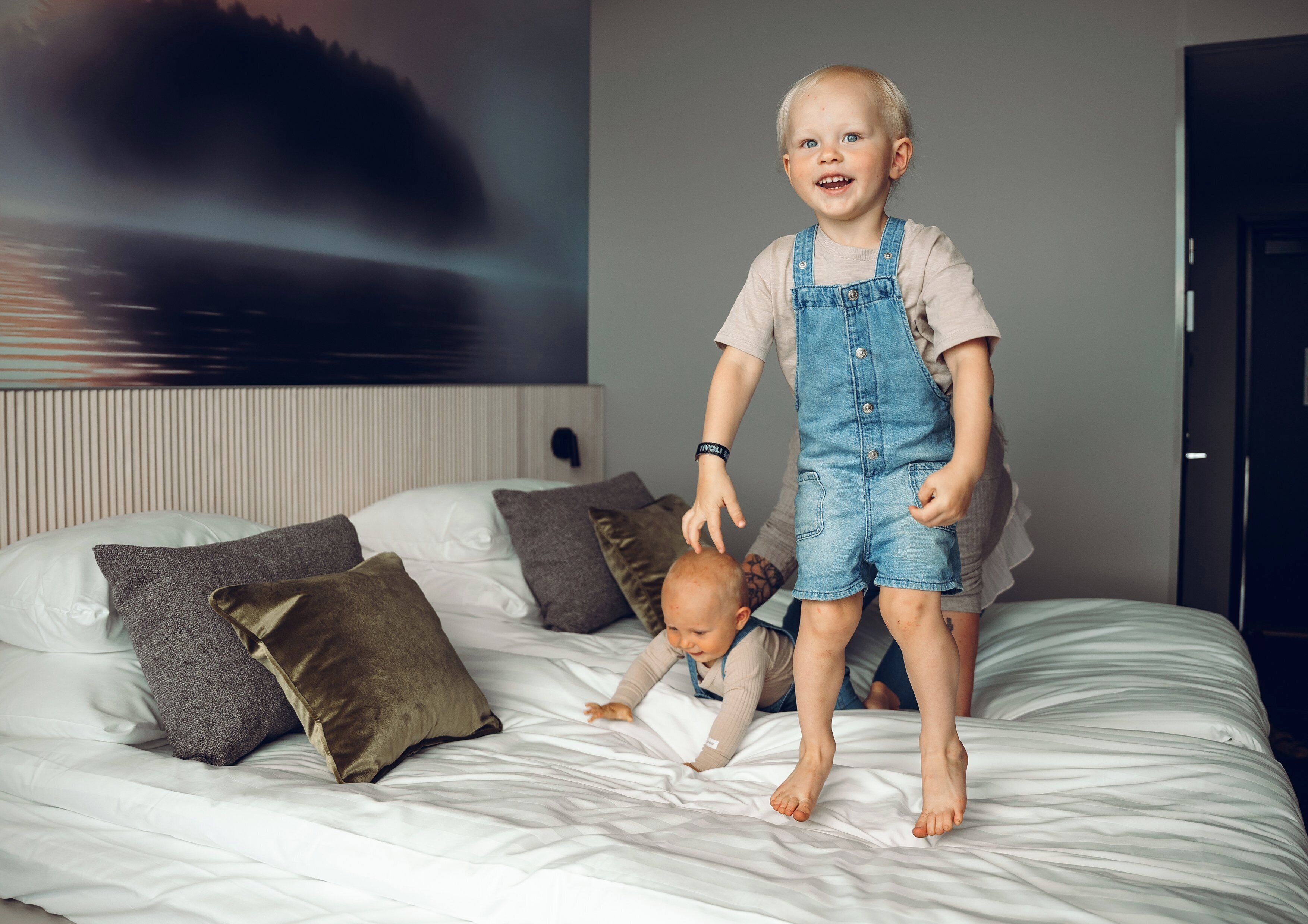 Un niño de pie sobre una cama, con un mural de un paisaje urbano en la pared de fondo.