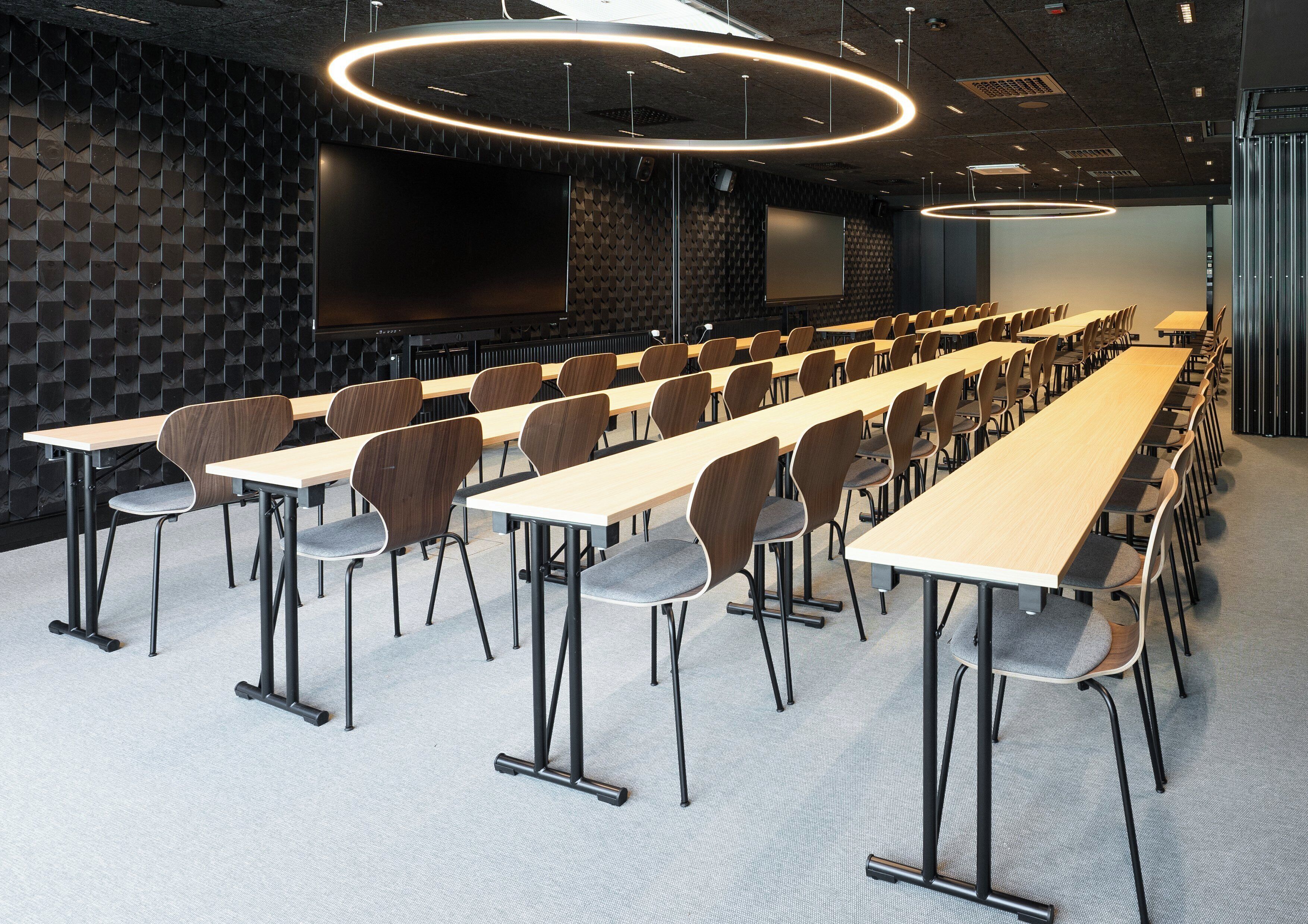 Ein Konferenzraum mit langen Tischen und Stühlen, einer Projektionsleinwand und einer Wand mit dunklem Muster.