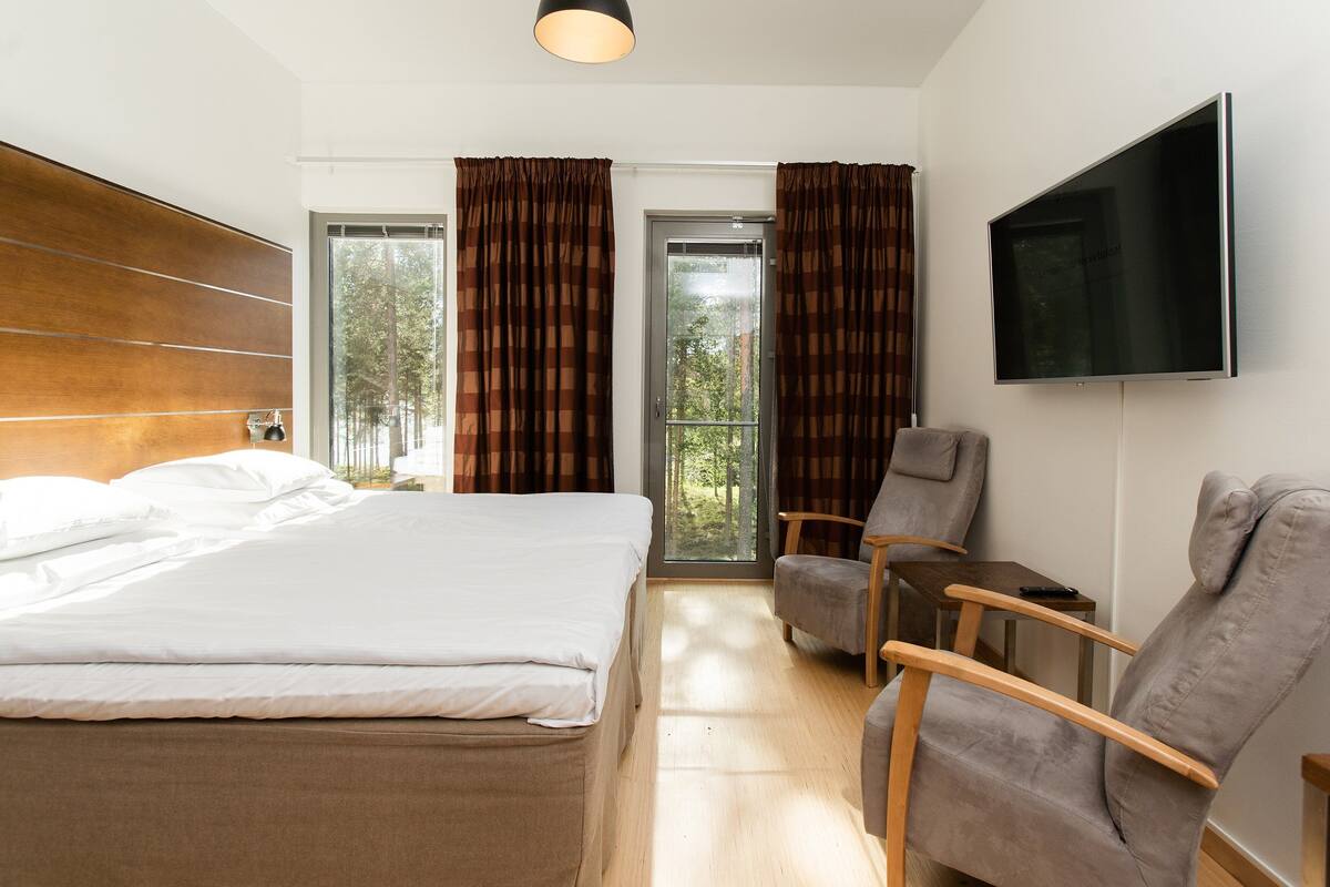 A modern hotel room with a large bed, a desk, a chair, a TV, and large windows with curtains.