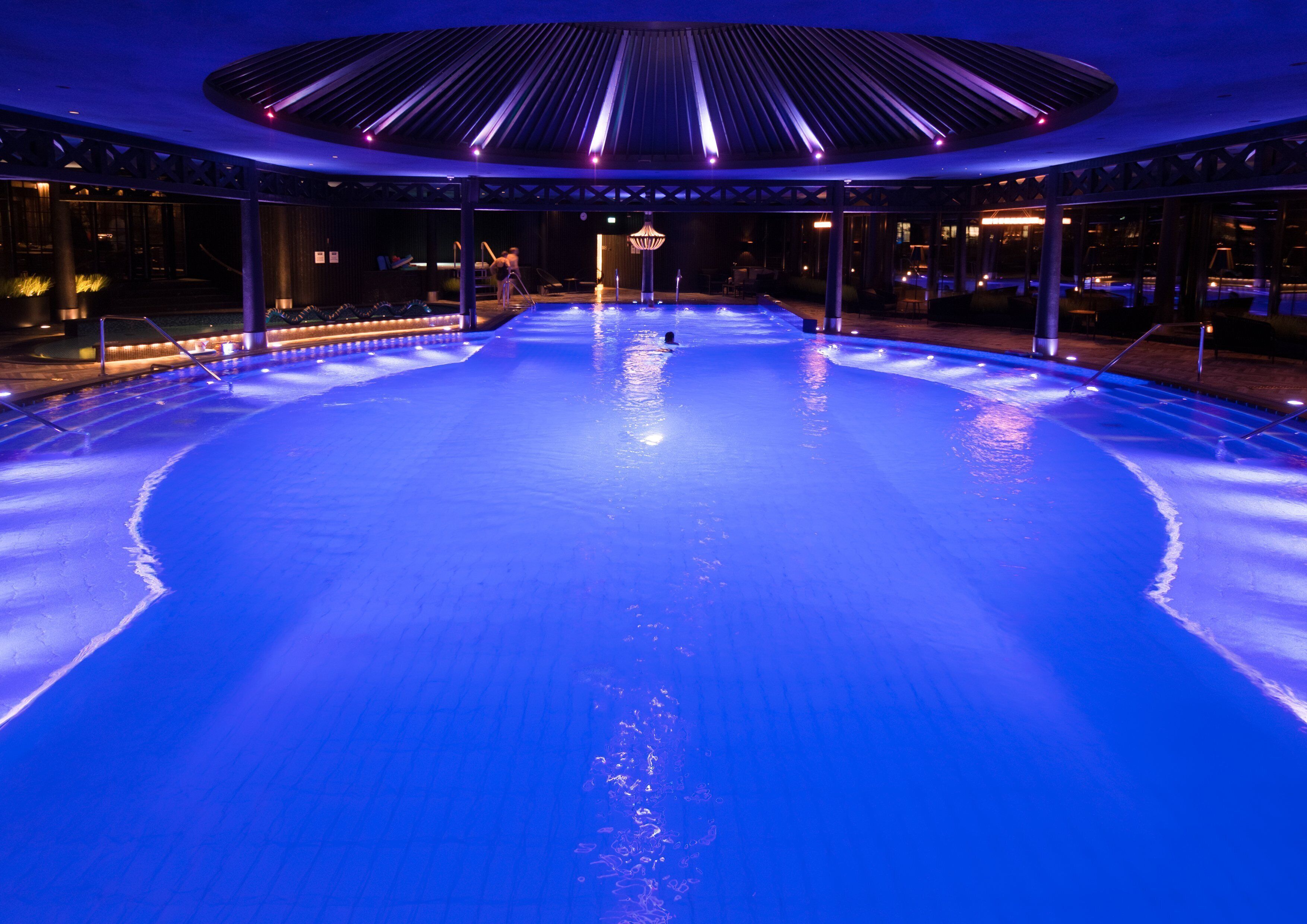 A large indoor swimming pool with a central island and surrounding seating areas, illuminated by blue lighting.