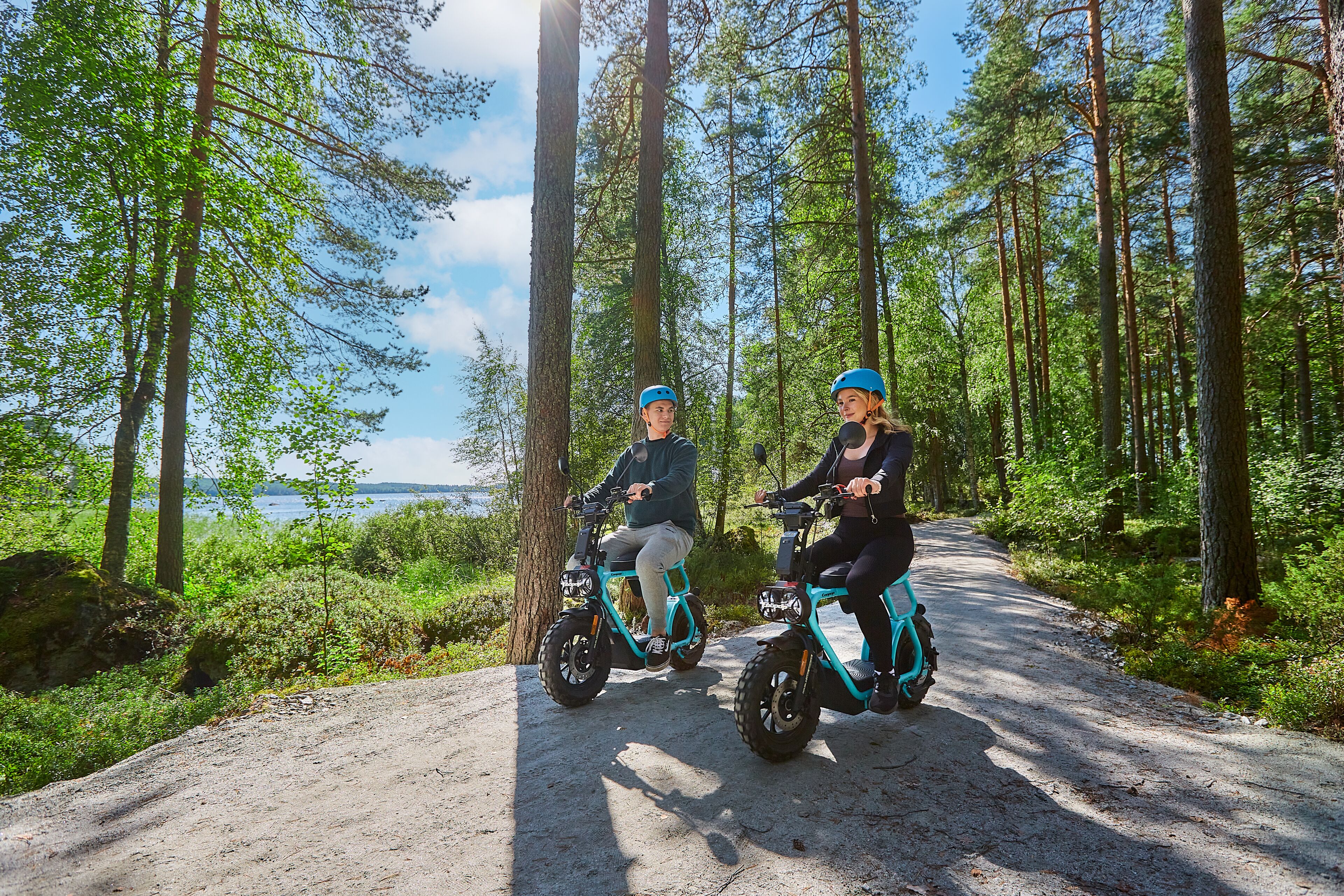 Deux personnes font du vélo électrique sur un chemin de terre, entourées d’arbres.