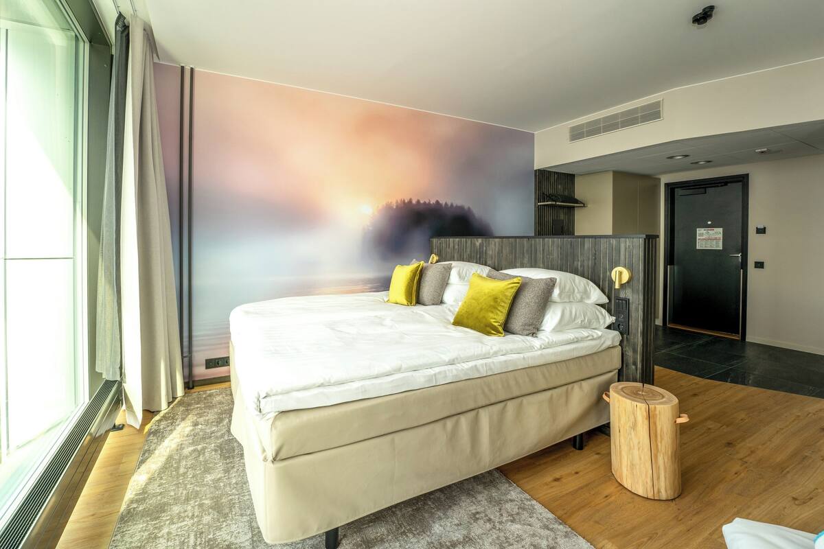 A modern hotel room with a large bed, a view of a forest through a window, and a wooden floor.