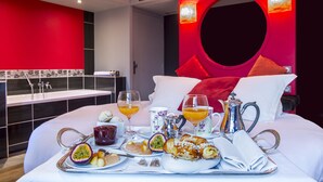 A breakfast setup on a bed with a red accent wall, a sink, and a modern bathroom in the background. - Carrion Cuisine Hôtel & Spa (Vire)