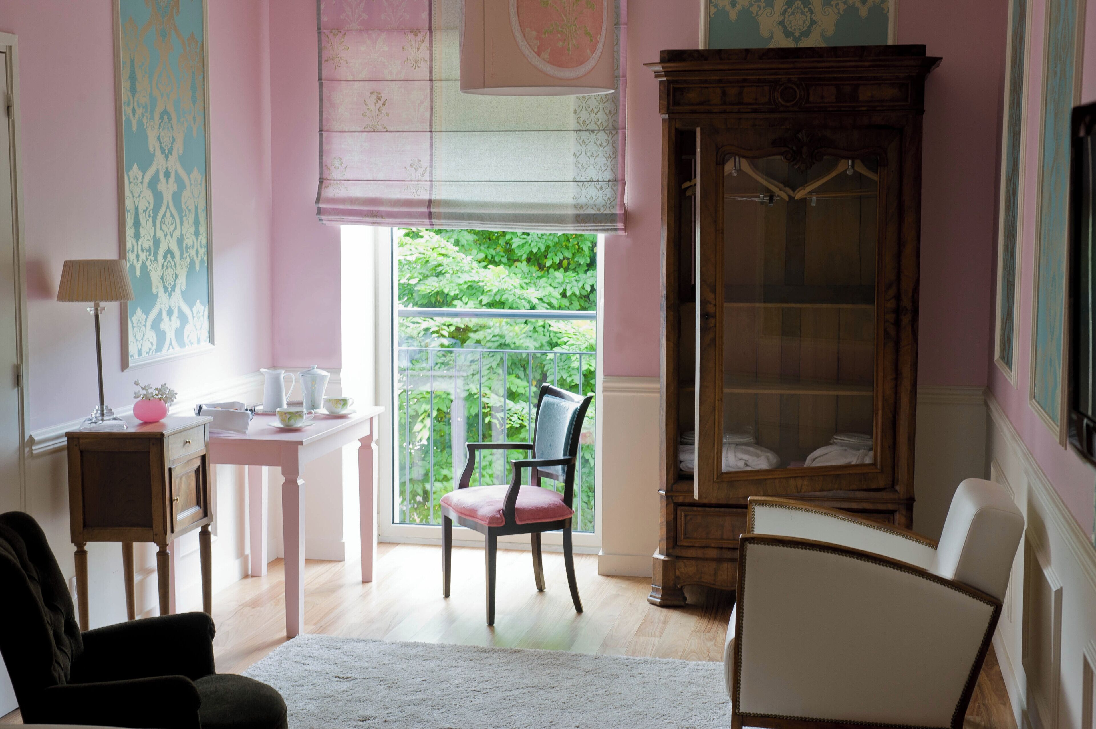 A room with a wooden cabinet, a chair, a table with a lamp, and a window with a view of greenery.