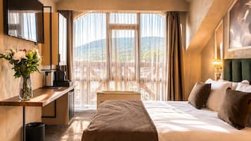 Habitación de hotel con cama, escritorio con cafetera, televisor, ventana con cortinas y balcón con vistas a las montañas.