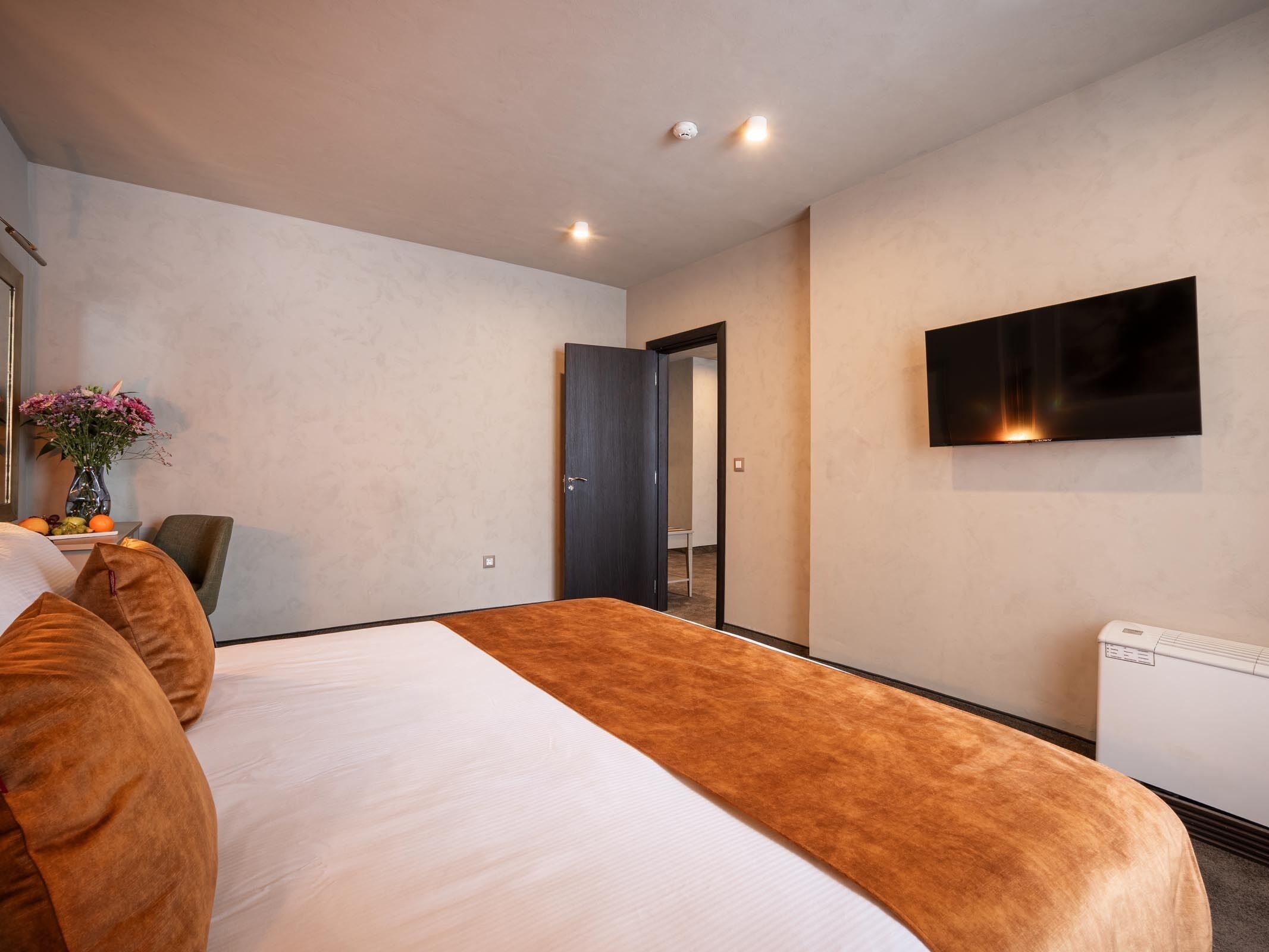 Une chambre d’hôtel moderne avec un grand lit, un téléviseur à écran plat fixé au mur et une petite table sur laquelle est posé un vase de fleurs.