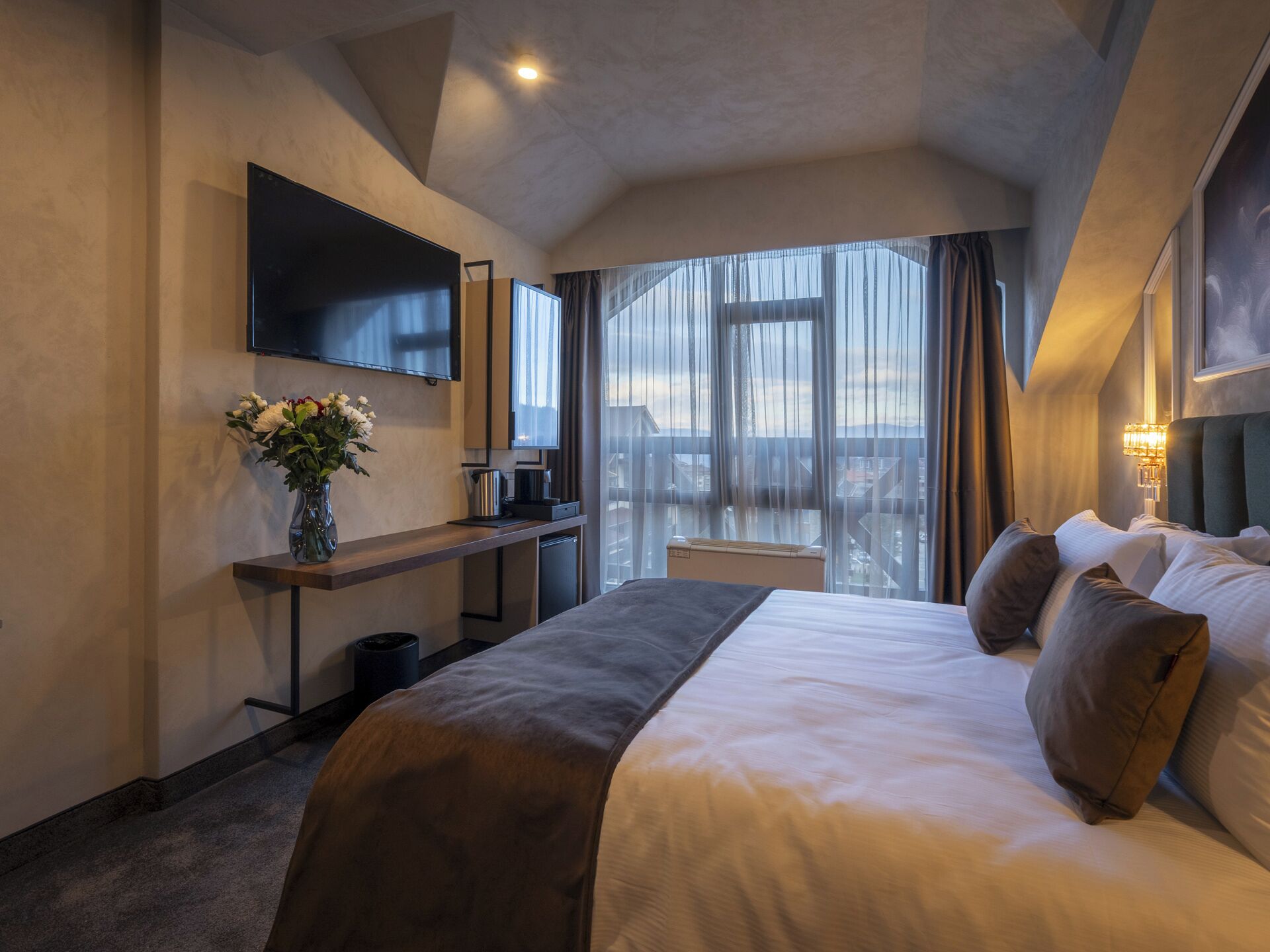 A modern hotel room with a large bed, a flat-screen TV mounted on the wall, a desk with a vase of flowers, and a view of the city through large windows.