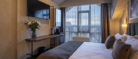 A modern hotel room with a large bed, a flat-screen TV mounted on the wall, a desk with a vase of flowers, and a view of the city through large windows.