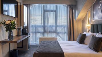 Un hotel moderno con una cama grande, un escritorio con cafetera, un jarrón de flores y vistas a la ciudad a través de cortinas translúcidas.