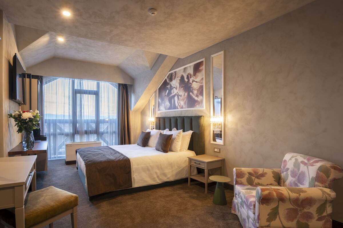 Ett hotellrum med en säng, ett skrivbord, en stol, ett litet bord och ett stort fönster med gardiner.