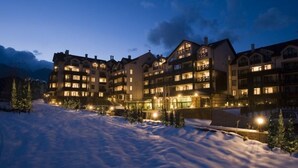 A multi-story hotel with a snow-covered ground, illuminated windows, and a mountainous backdrop.