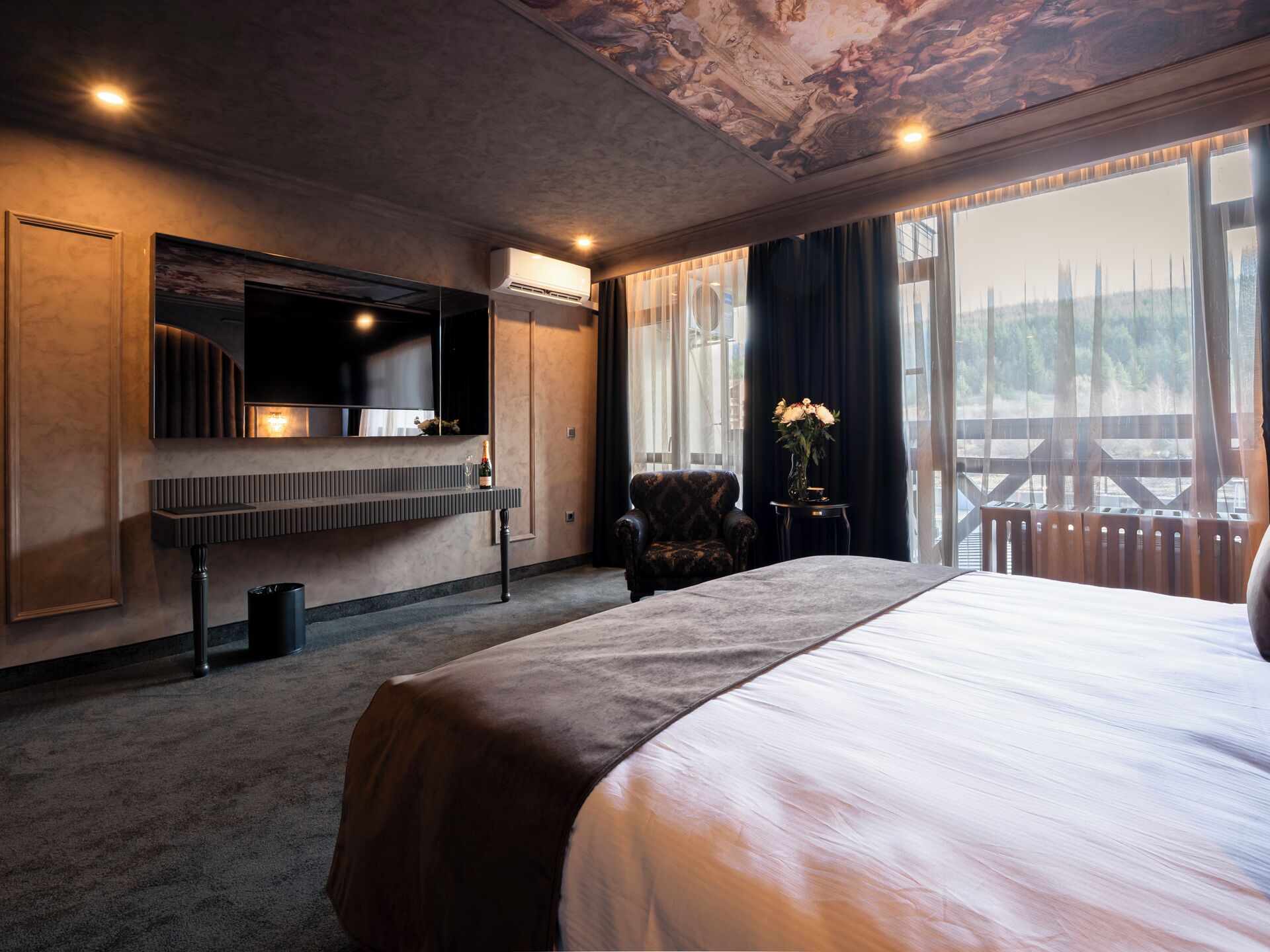 A hotel room with a large bed, a flat-screen TV mounted on the wall, a small side table with a vase of flowers, and a view of a wooden deck through large windows.