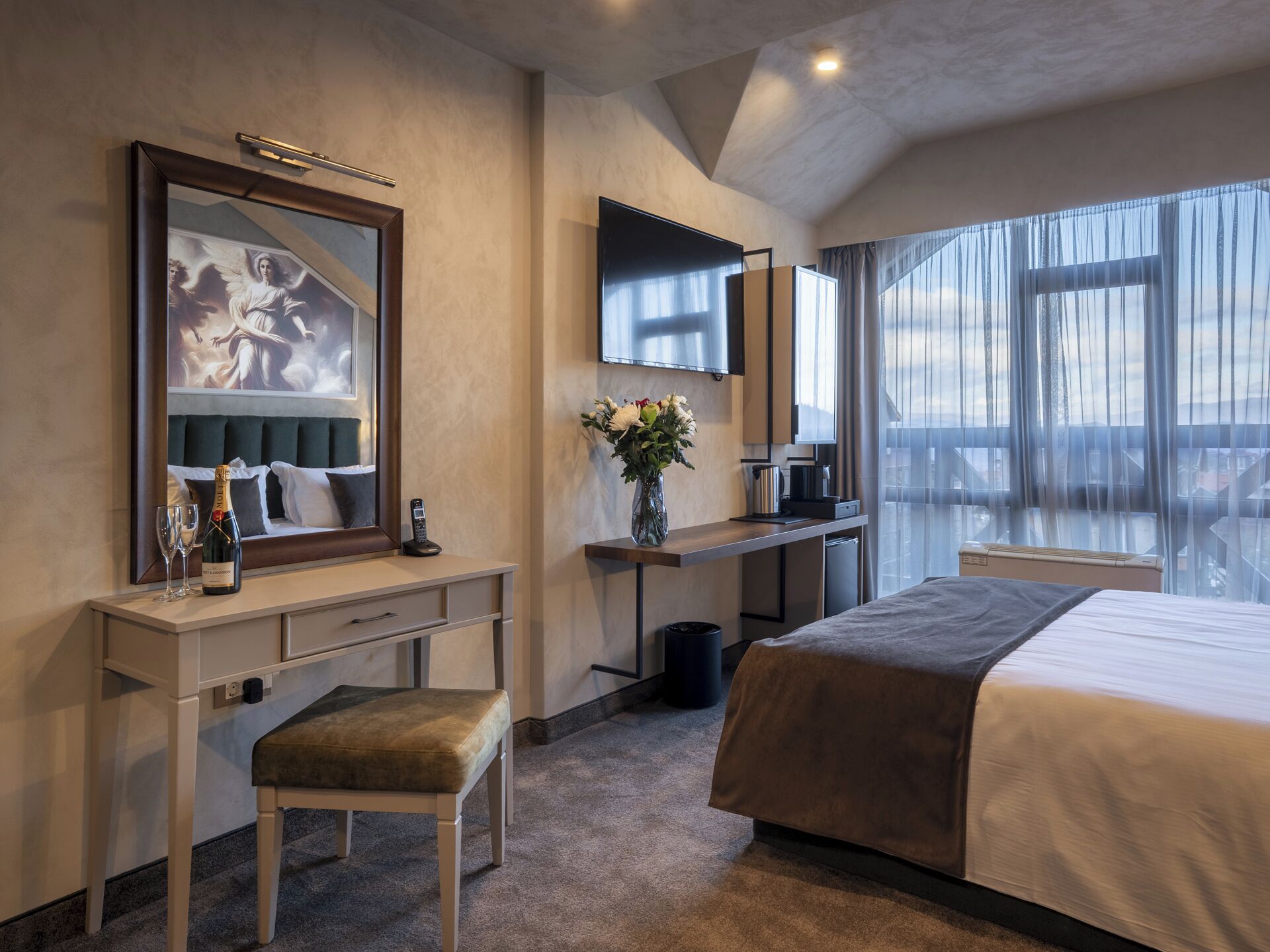 A hotel room with a bed, a desk with a chair, a TV, a window with curtains, and a painting on the wall.